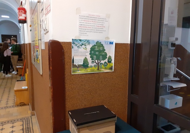 Szkolny korytarz z plakatem promującym naukę języków oraz pudełkiem do wrzucania odpowiedzi w konkursie